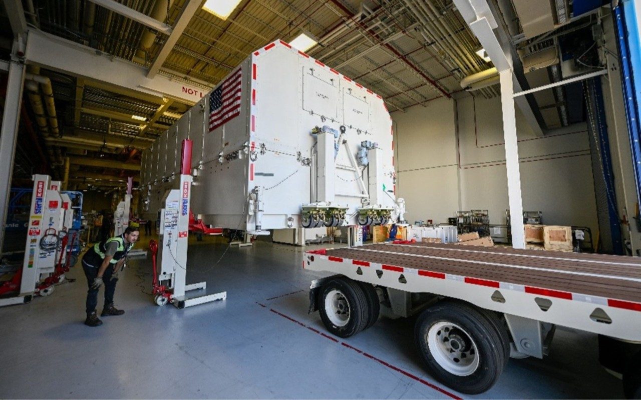 a male Lockheed Martin technician looks at a truck that is being loading with the GPS III SV07 satellite payload